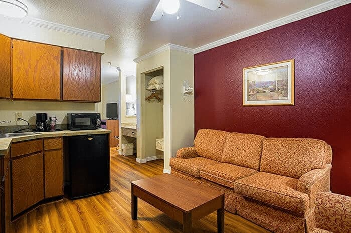 Cozy hotel room interior featuring a kitchenette with wooden cabinets, a black refrigerator, a coffee maker, a patterned sofa, and a wooden coffee table against a maroon accent wall, showcasing Listing Marketing Pros' hotel photography expertise.