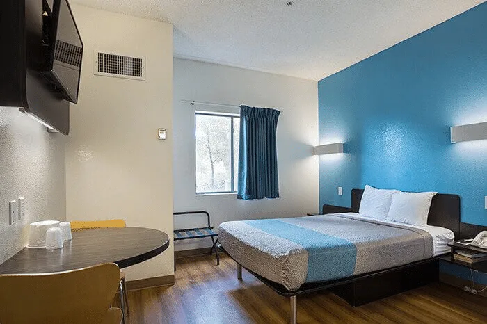 Hotel room featuring a bed with blue accent wall, table and chairs, window with blue curtains, and flat-screen TV, showcasing Listing Marketing Pros' hotel portfolio.